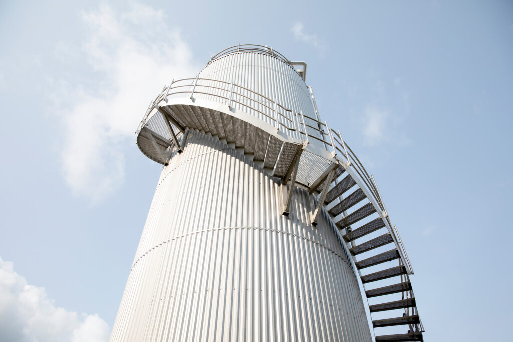 A cage ladder attached to a large stainless steel storage tank, set against a clear blue sky, highlighting industrial safety and access features.